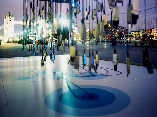 Phones hanging on cables inside a glass tank on a London riverside at night, with Tower Bridge behind